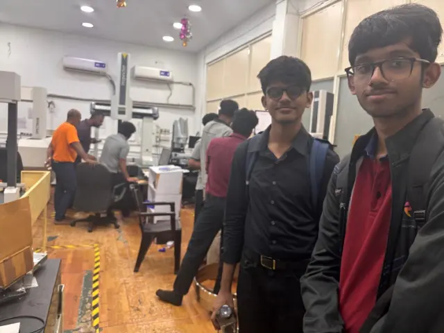 Adithya and a teammate in a machine shop, a CNC machine and a coordinate measuring machine behind them