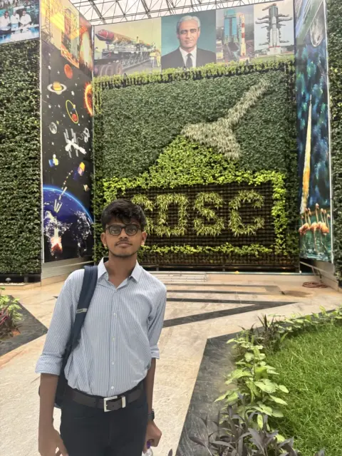 Adithya in front of the SDSC sign in a wall of plants at Satish Dhawan Space Centre