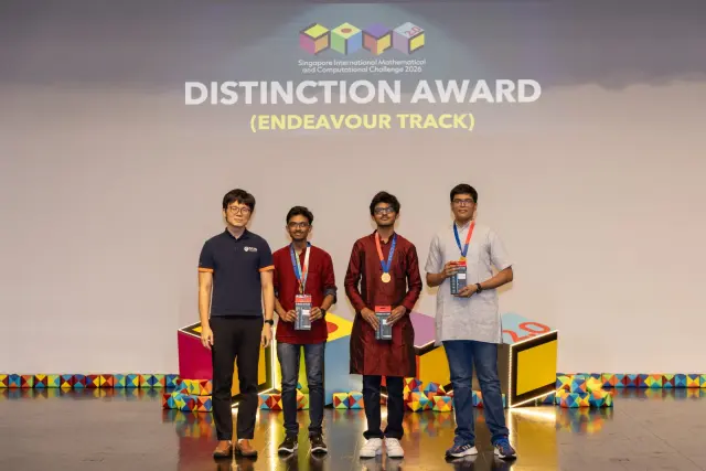 The team of three on stage under the Distinction Award, Endeavour Track banner, with a presenter, at SIMC 2026